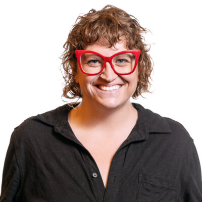 Ally Mills smiling headshot wearing a black button down shirt and big red glasses