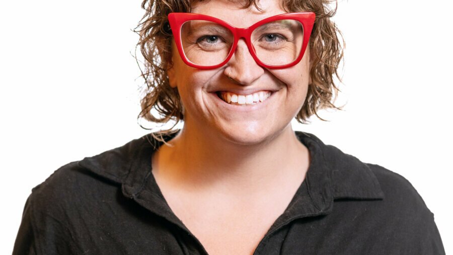 Ally Mills smiling headshot wearing a black button down shirt and big red glasses