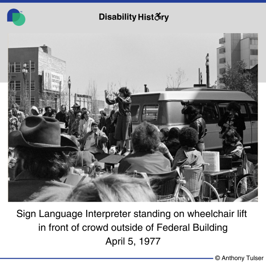 Black and white photo of crowd of protesters outside of the Federal Building. A sign language interpreter stands on a van wheelchair lift, signing to the crowd. Text: Sign Language Interpreter standing on wheelchair lift in front of crowd outside Federal Building. April 5. Source: Anthony Tusler. 