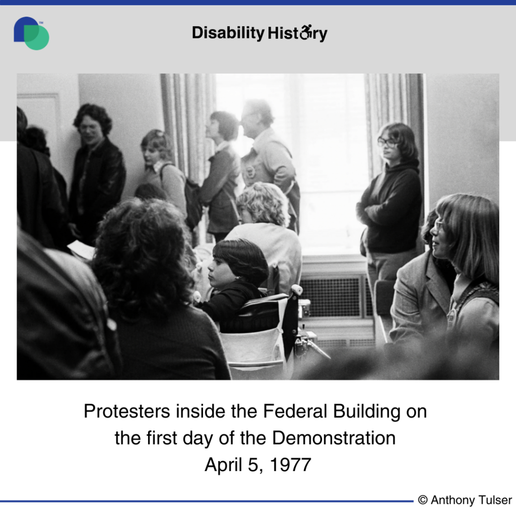 Black and white photo of protesters sitting and standing inside the Federal Building in front of a window. Text: Protesters inside the Federal Building on the first day of the demonstration. April 5. Source: Anthony Tusler. 