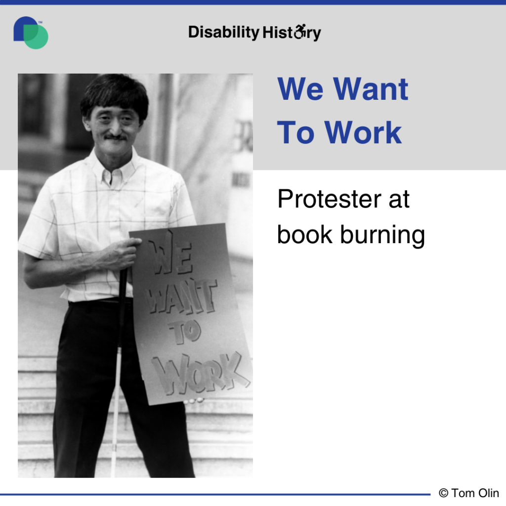 Black and white photo of man with a mustache smiling while holding his guide cane and showing a sign that says, “We Want to Work.” Text: We Want To Work. Protester at book burning. Source: Tom Olin.