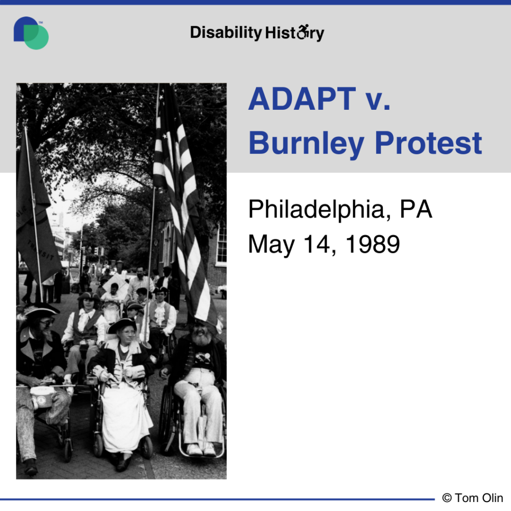 Black and white photo of group of protesters in wheelchairs marching down a sidewalk wearing revolutionary attire. One protester is holding the ADAPT American flag. Text: ADAPT v. Burnley Protest. Philadelphia, PA. May 14, 1989. Source: Tom Olin.