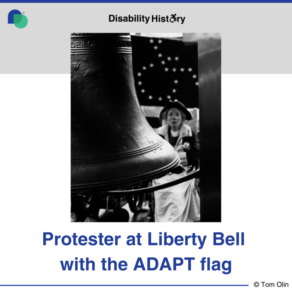Black and white photo showing part of the liberty bell. Behind the bell is a protester in revolutionary attire in a wheelchair with the ADAPT American Flag behind her. Text: Protester at Liberty Bell with ADAPT flag. Source: Tom Olin.