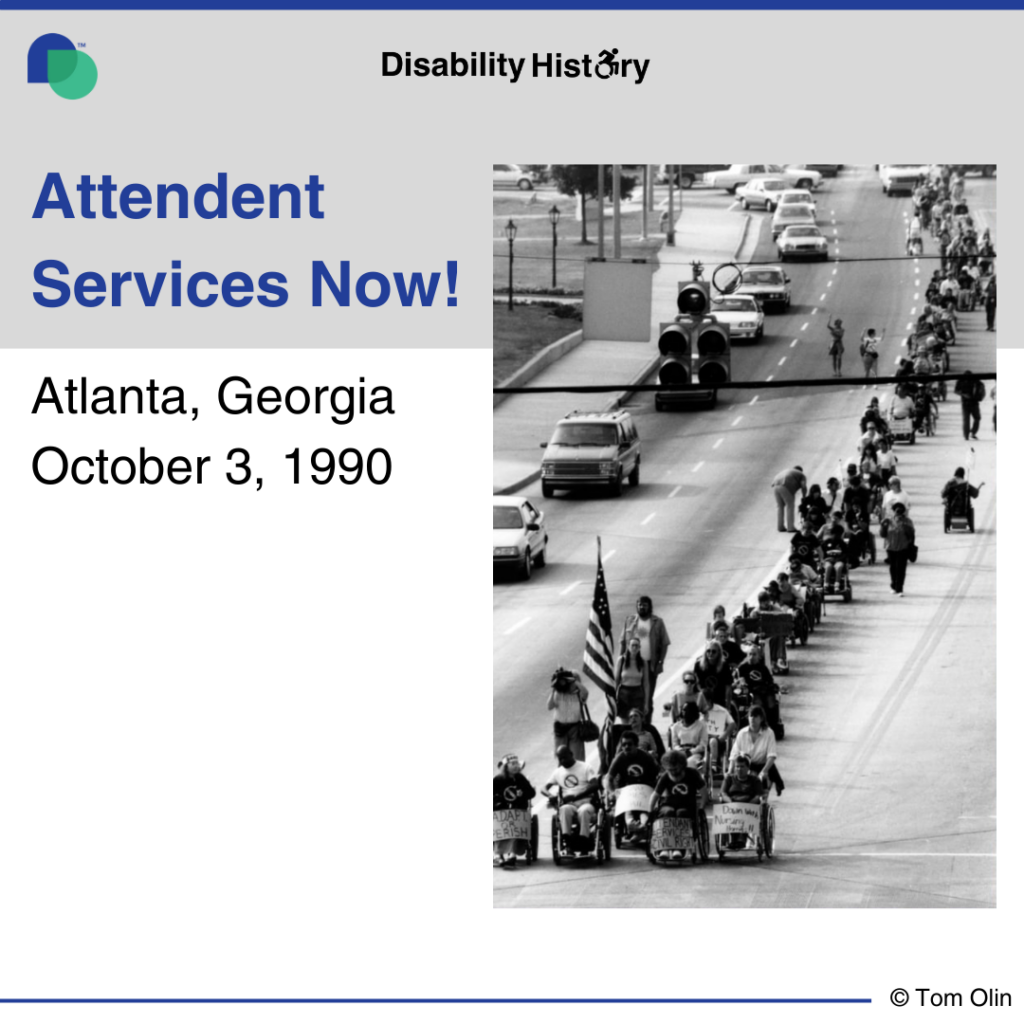 Black and white photo of line of protesters, many in wheelchairs, marching up one side of the road with cars on the other side. One protester in the front of the line holds the ADAPT American flag.