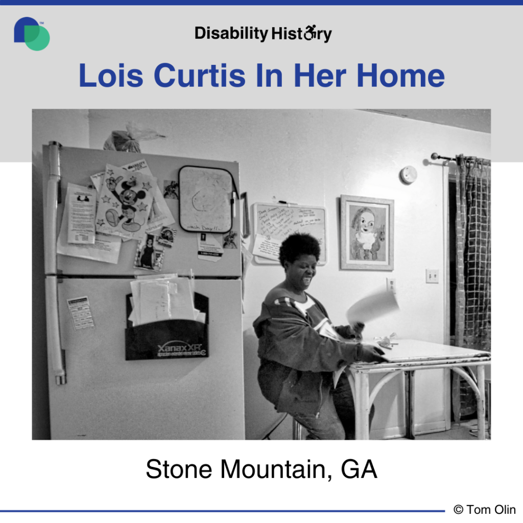 Black and white photo of Lois Curtis sitting at her kitchen table, smiling with a paper in her hand. To the left of her is her fridge and behind her is a white board and framed drawing. Text: Lois Curtis in her home. Stone Mountain, GA. Source: Tom Olin