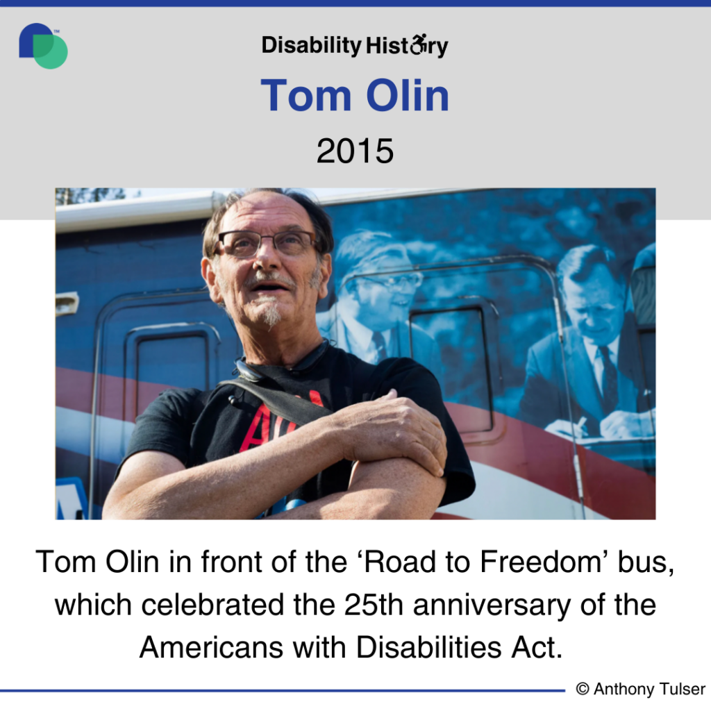 Color photo of Tom Olin standing with his arms crossed in front of the ‘Road to Freedom’ bus that has the American flag on it and Tom’s photo of George H.W. Bush signing the ADA. Text: Tom Olin in front of the Road to Freedom bus, which celebrated the 25th Anniversary of the Americans with Disabilities Act. Source: Anthony Tusler.