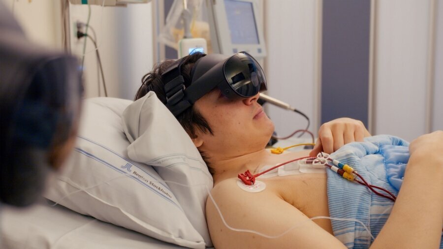 A person wearing a virtual reality headset lying in a hospital bed with medical equipment around them in a still from The Reality of Hope