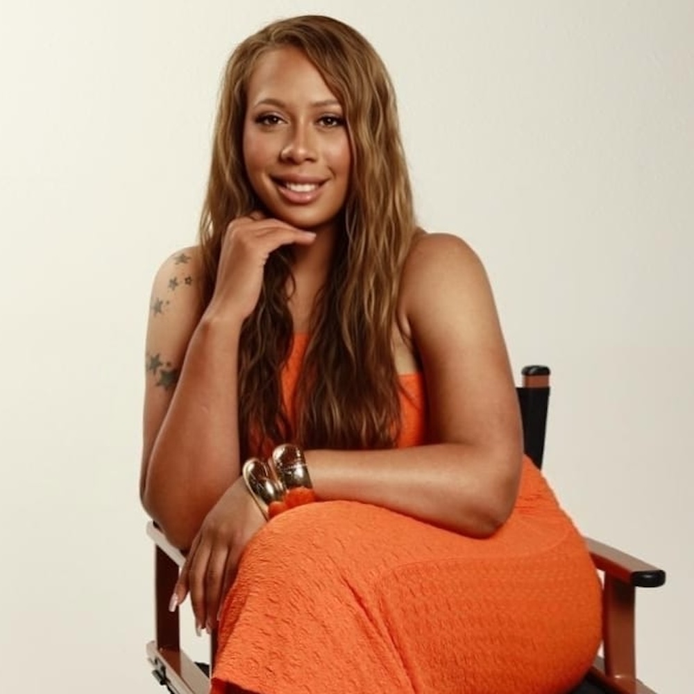 Charlie T Savage headshot wearing an orange dress seated in a chair