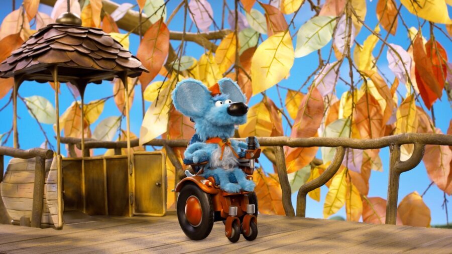 still of Jeff Mouse from Donkey Hodie, a blue mouse puppet who uses a wheelchair. there are orange and yellow leaves in the background behind Jeff.