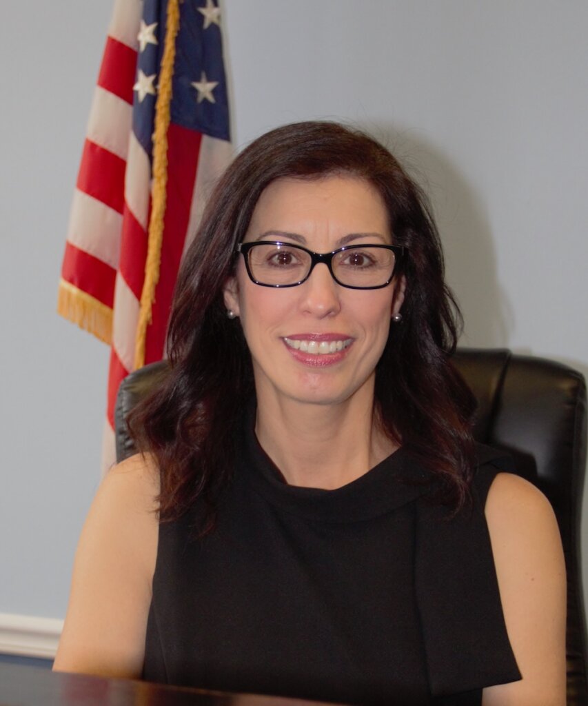 Dina Grilo smiling headshot seated in front of an American flag
